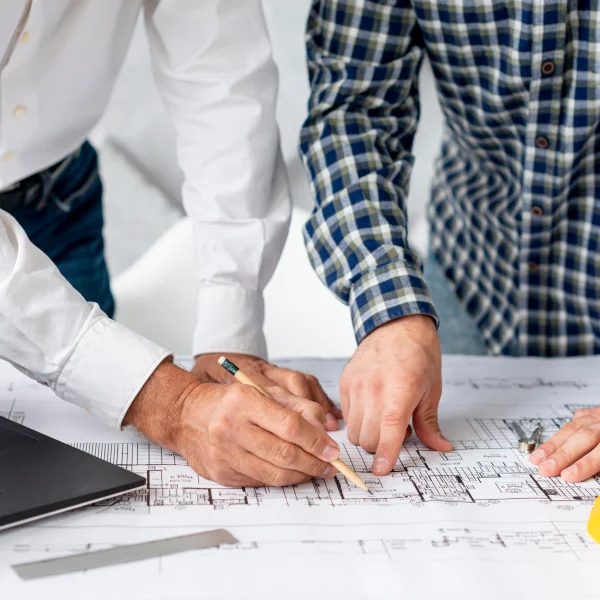 Two men collaborating on a construction blueprint while using a laptop for project planning.