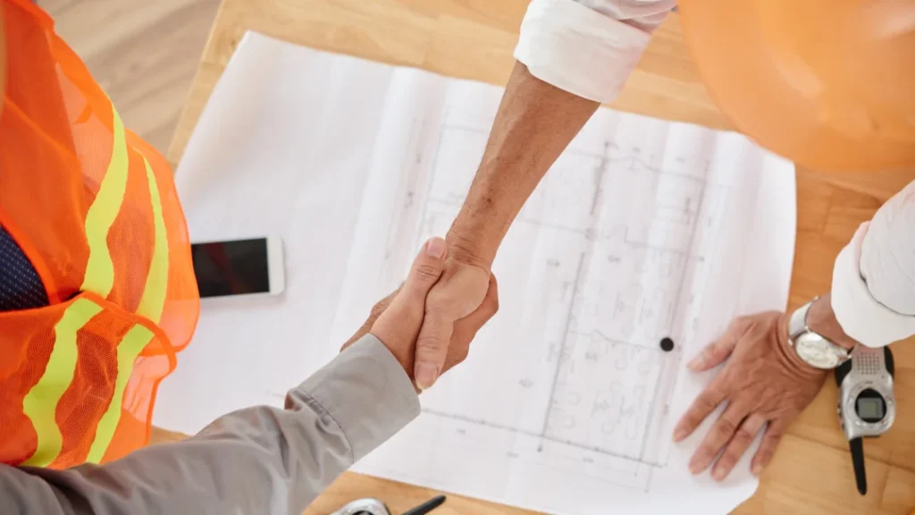 Two people shaking hands over a blueprint, discussing plans for a renovation project.